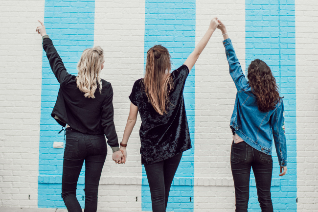 three women holding hands facing a blue and white striped building