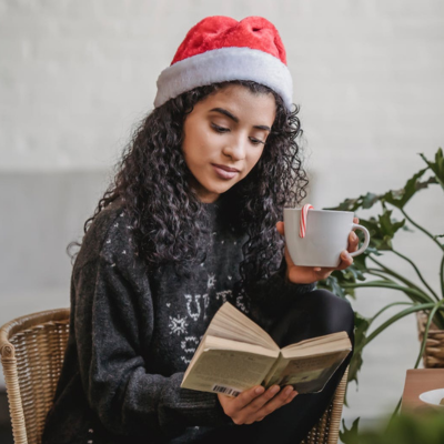 woman wearing Santa hat reading a book and holding cup of cocoa