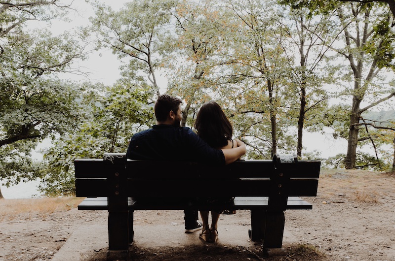 man looking to woman sitting on a bench in front of trees and a lake