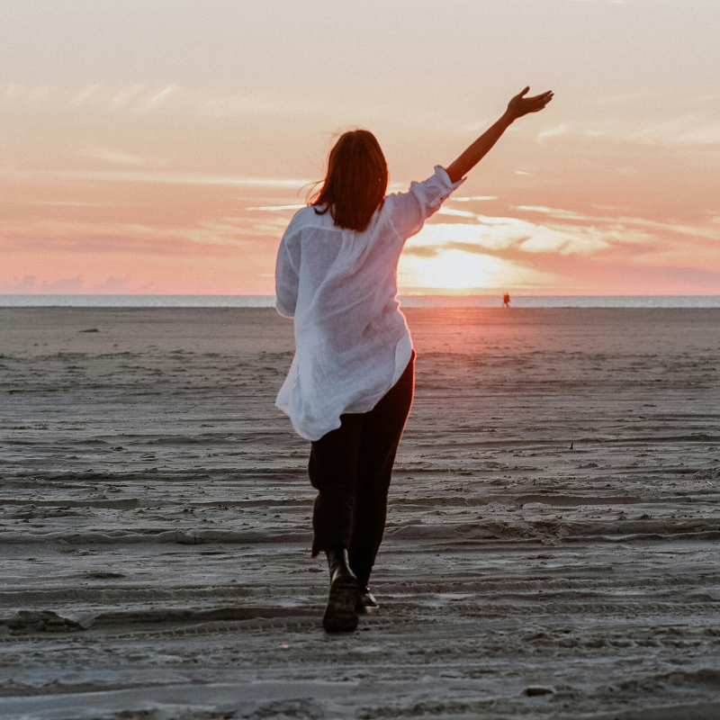 woman standing on a sandy beach at sunset with her arm outstretched